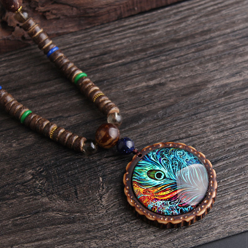 pendentif plume de paon ethnique posé sur bois brut – chaîne de perles et breloque dorée visible