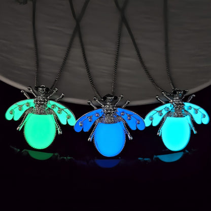 Trois colliers pendentif insecte phosphorescents côte à côte dans le noir — lueurs verte, bleue et turquoise, comparatif des 3 variantes de couleur